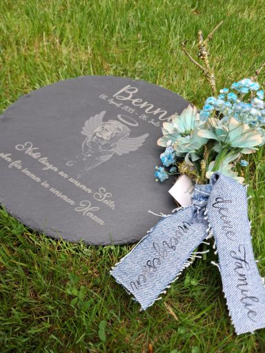 Gedenktafel mit floralen Elementen und blauem Band auf grasigem Untergrund.
