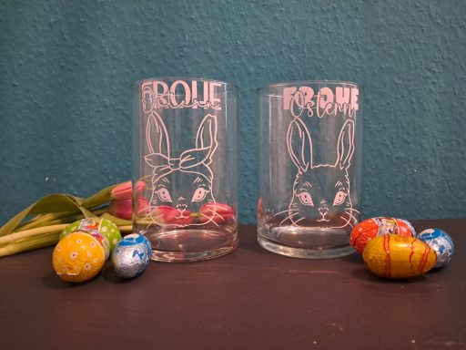 Zwei Gläser mit Hase-Design, daneben bunte Ostereier und eine Blume.